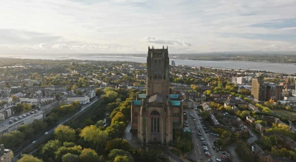 Aerial view of Liverpool Cathedral overlooking Liverpool with surrounding neighborhoods, greenery, and the River Mersey under a soft cloudy sky
