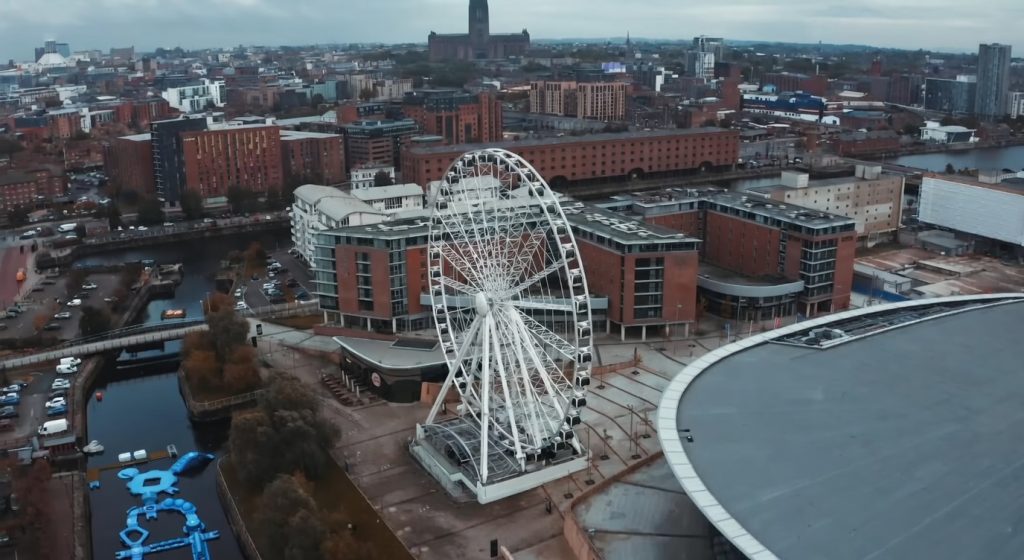Alt text (SEO-friendly):“Aerial view of Tower Bridge spanning the River Thames in London, with historic stone towers, blue suspension elements, and city skyline under a cloudy sky.”
