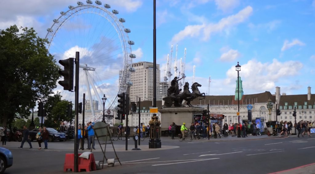 London street scene featuring the London Eye and Boudica Statue near Westminster Bridge, with pedestrians, traffic lights, and city skyline under a blue sky
